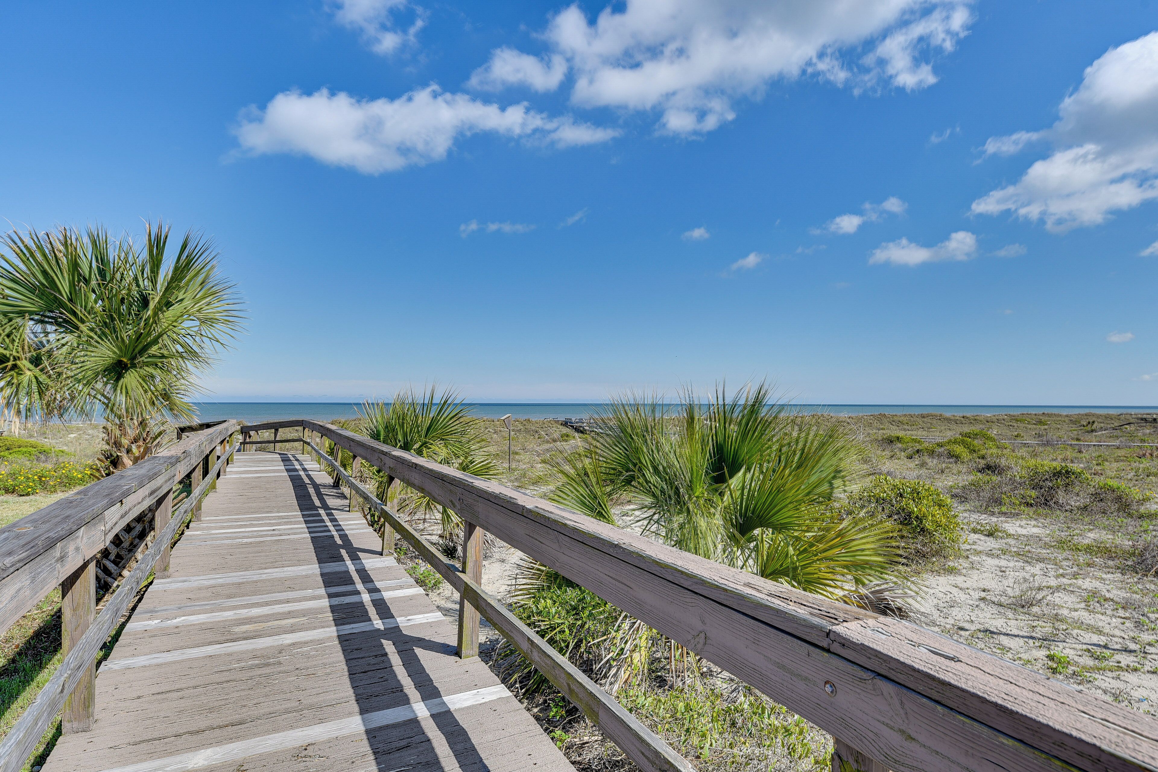 Une promenade en bois menant à la plage, bordée de palmiers et de dunes de sable.