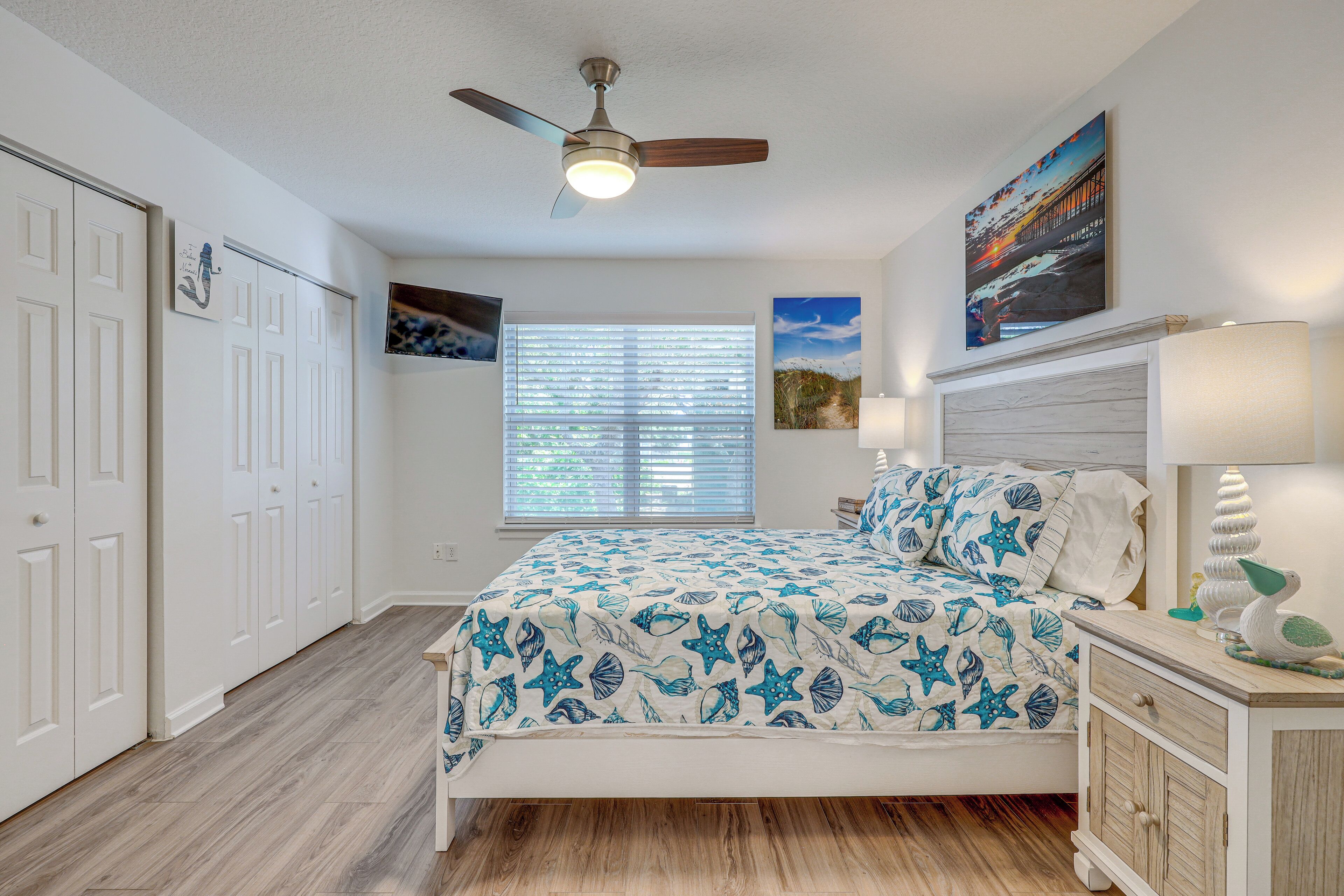 A bedroom with a bed, a ceiling fan, a window with blinds, a nightstand, and a wall with artwork.