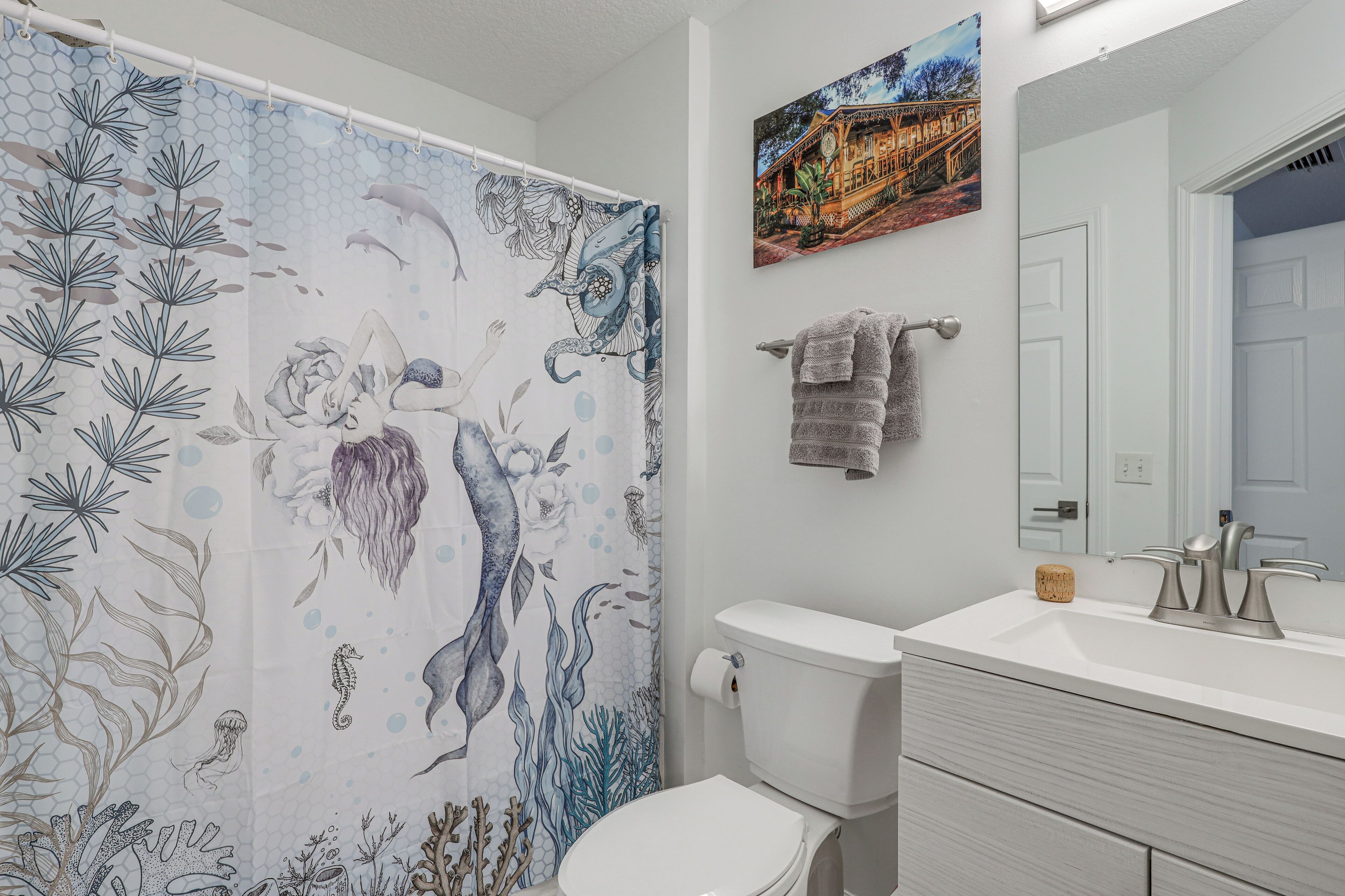 A bathroom with a shower curtain featuring a mermaid and sea creatures design, a toilet, a sink with a faucet, a towel rack with a towel, and a framed picture on the wall.