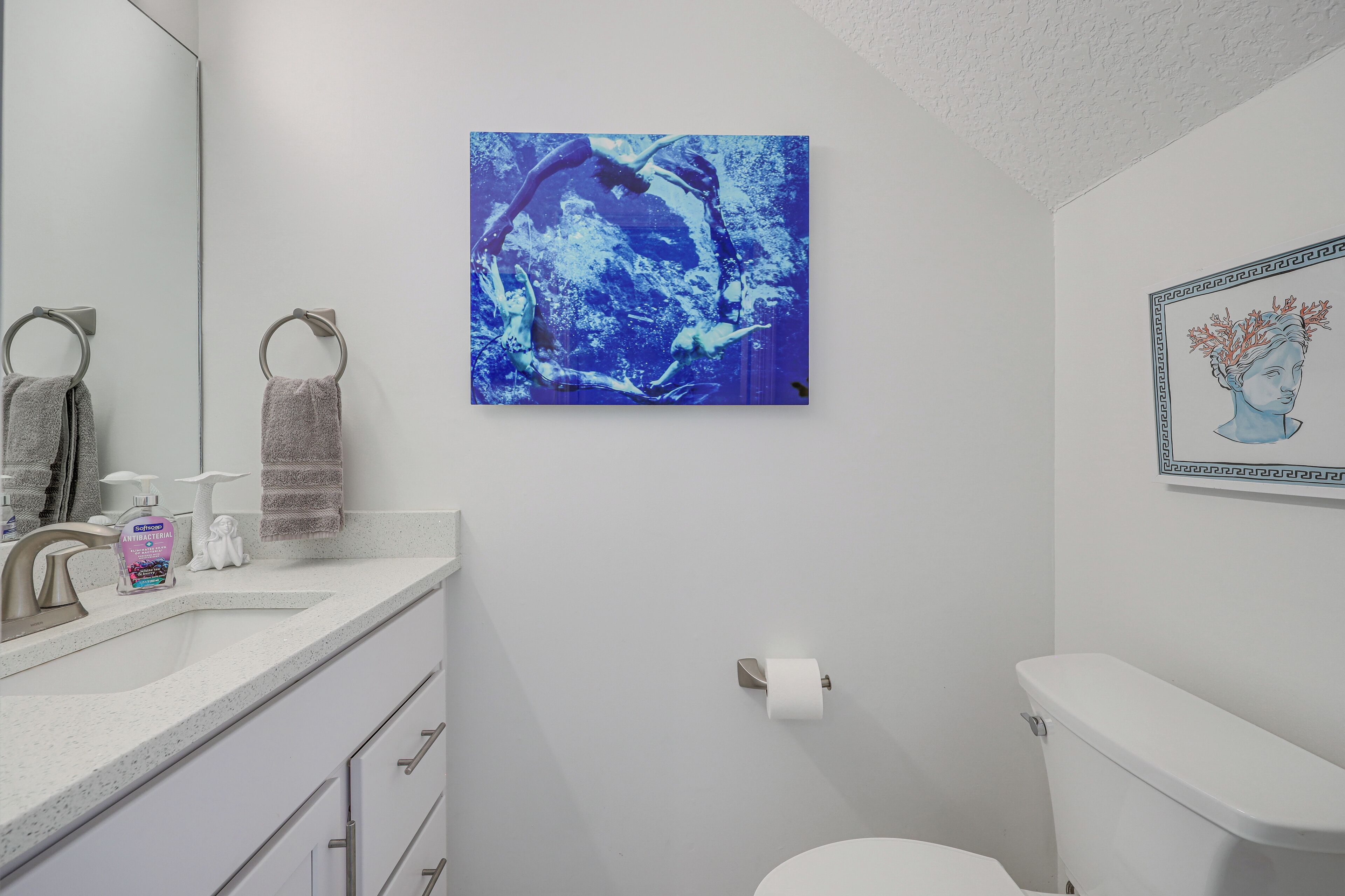 A bathroom with a white sink, a mirror, a toilet, and a blue artwork on the wall.