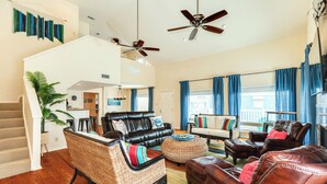 A spacious living room with a leather sofa, a wicker chair, and a brown leather armchair. There are two ceiling fans, a staircase, and a view of the city through large windows with blue curtains.