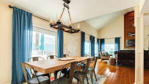 A dining area with a wooden table and metal chairs, a chandelier, and a view of the outdoors through large windows with blue curtains.