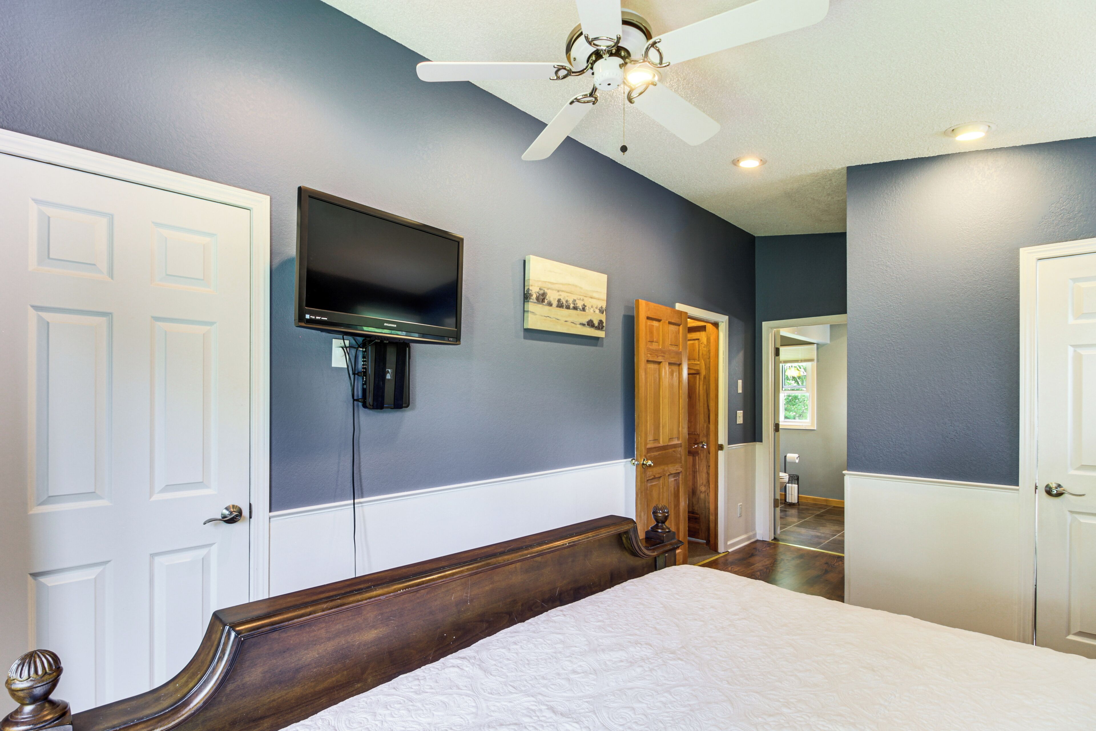 A bedroom with a bed, a ceiling fan, a mounted TV, and a door leading to another room.