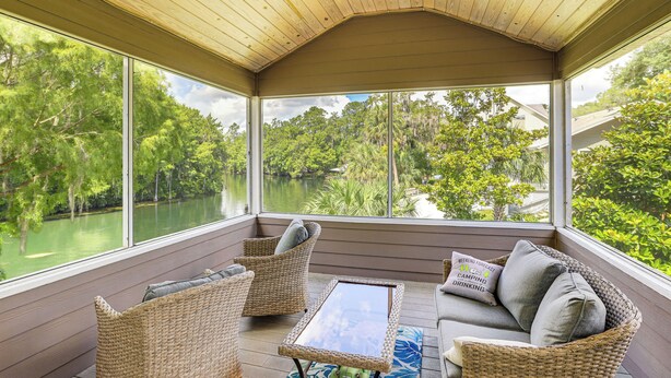 A covered porch with wicker furniture, a ceiling fan, and a view of a lake surrounded by trees. - Boathouse & Dock: Riverfront Dunnellon Getaway! (Dunnellon)