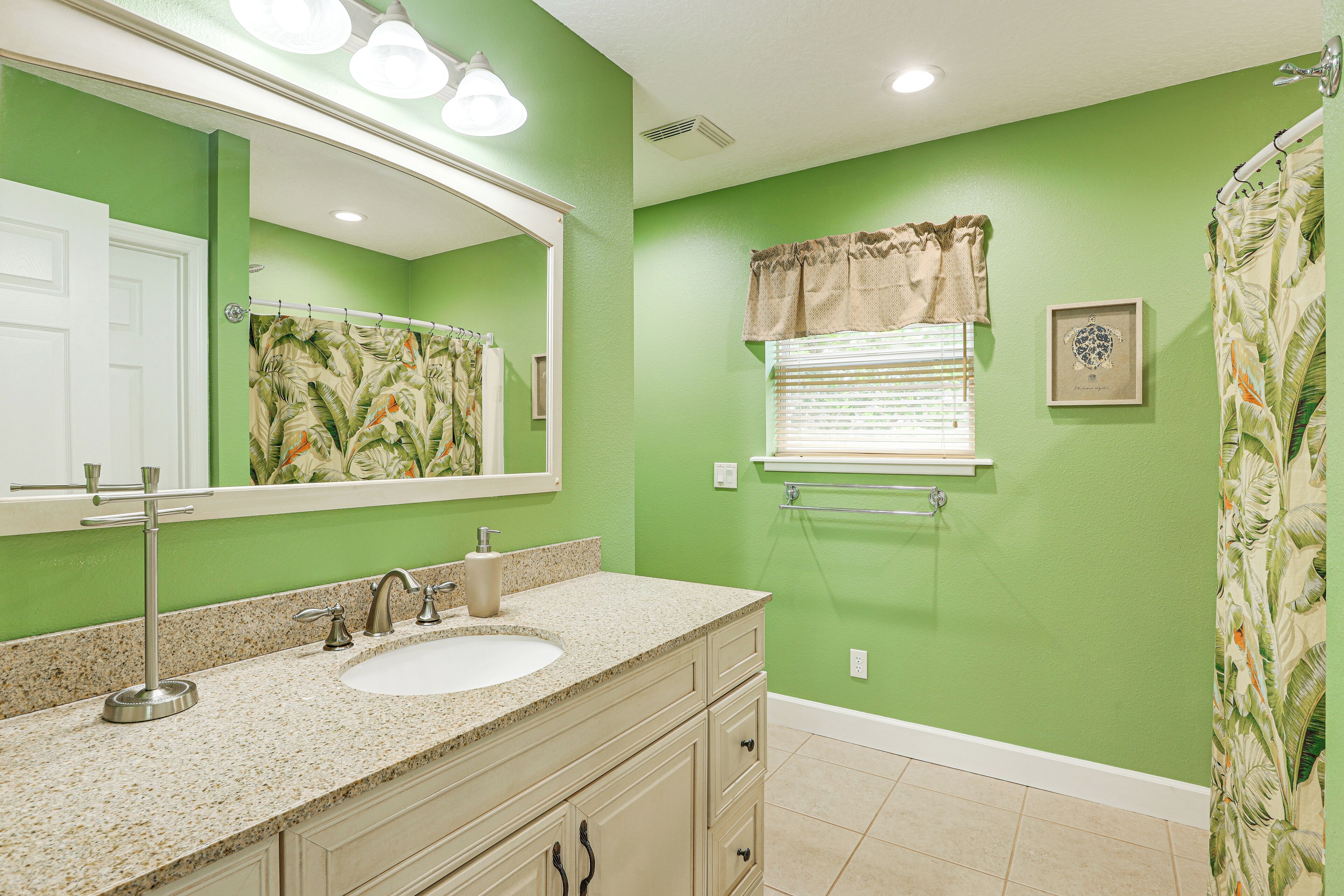 Une salle de bain avec un mur vert, un lavabo avec un meuble-vasque, un rideau de douche à motifs tropicaux et une fenêtre avec des stores.