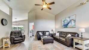 A spacious living room with a large brown leather sofa, a matching ottoman, and a wooden coffee table. A ceiling fan and a chandelier are visible.