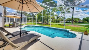 A covered outdoor swimming pool with lounge chairs and umbrellas, surrounded by a white fence and greenery.