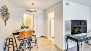 A modern kitchen with a dining area, featuring a wooden table, bar stools, a hanging light fixture, and a large flat-screen TV on a stand. - Dallas Condo in Walkable Knox Street District! (Dallas)