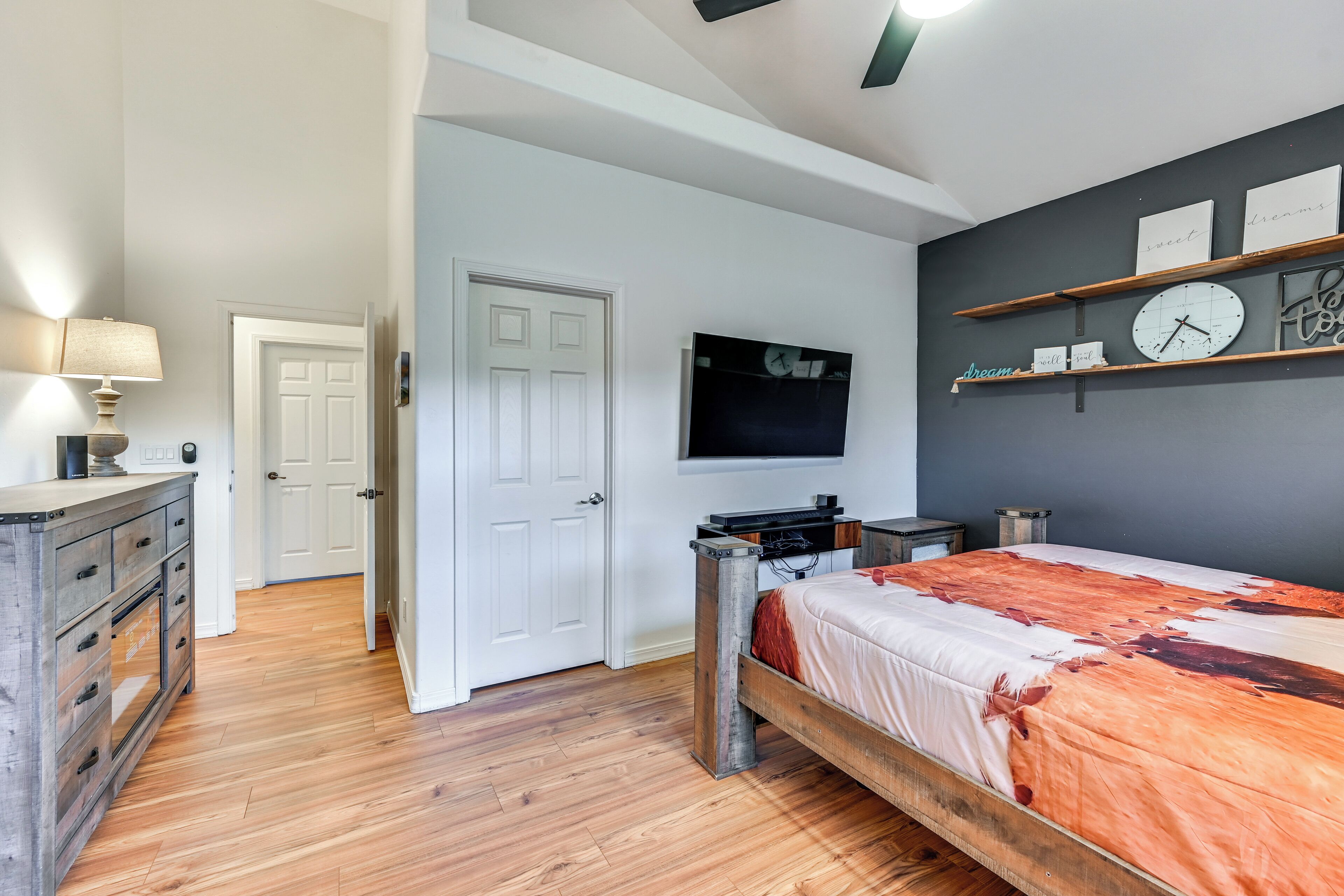 Un dormitorio con una cama de madera, un televisor montado en la pared, un aparador y una puerta que da a otra habitación.