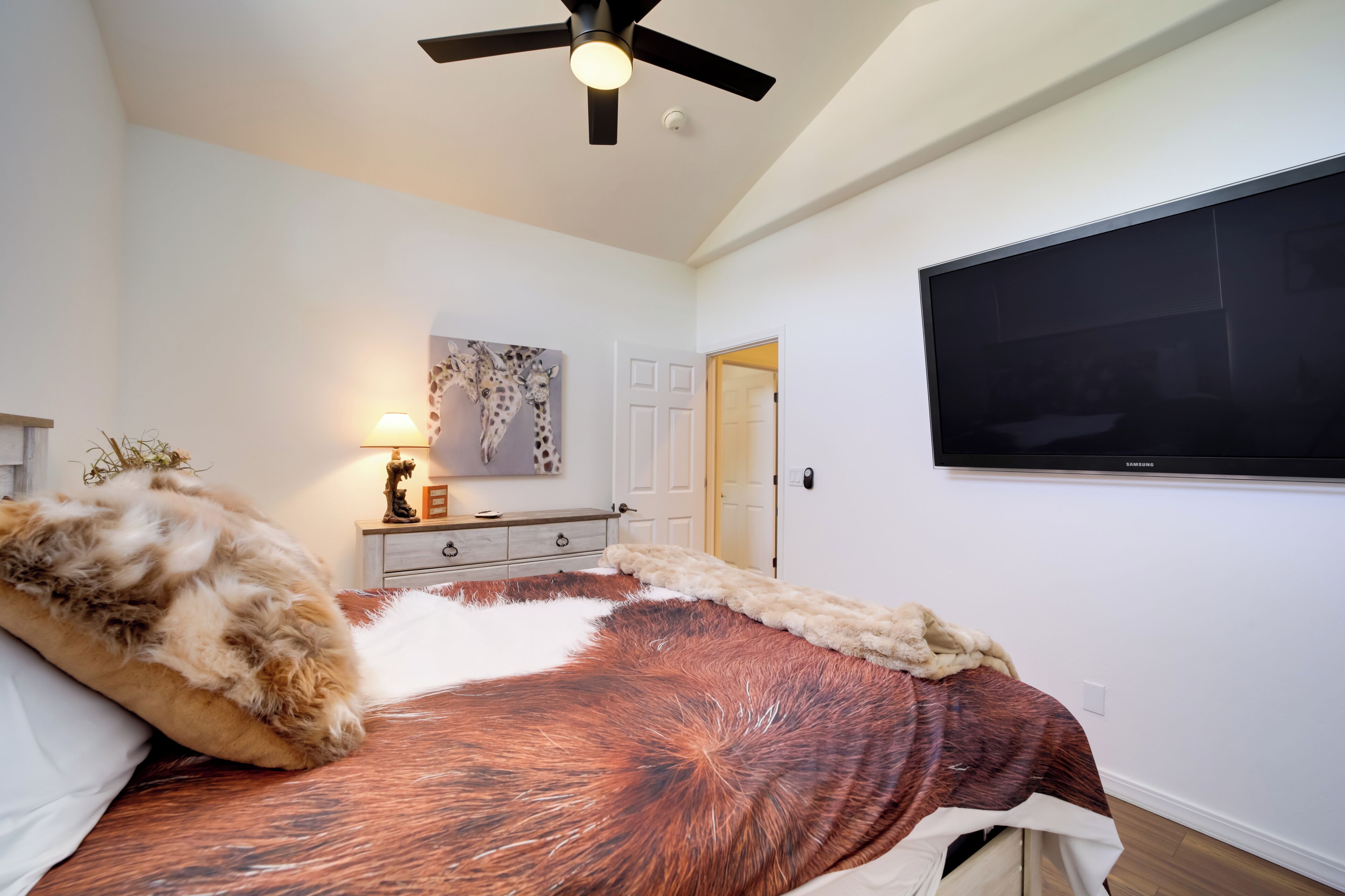 A bedroom with a bed, a ceiling fan, a TV mounted on the wall, and a door leading to another room.
