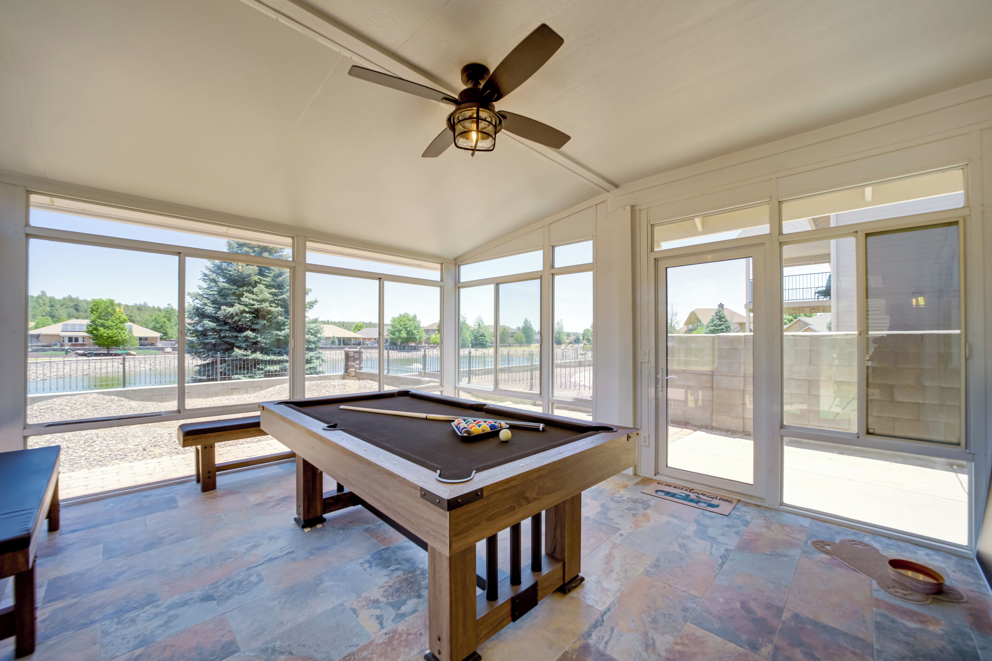 Une table de billard dans une pièce dotée de grandes fenêtres, d’un ventilateur de plafond et offrant une vue sur l’extérieur.