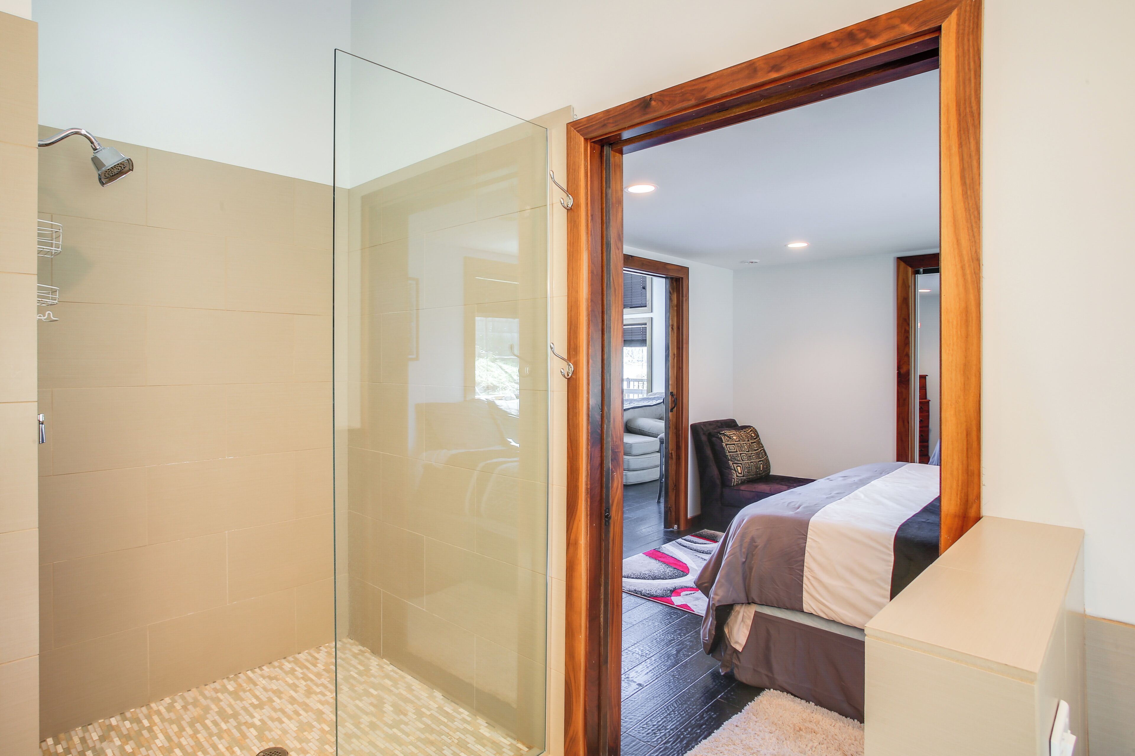 Ein modernes Badezimmer mit einer Duschkabine aus Glas und einer Holztür, die in einen Schlafbereich mit Bett und kleinem Tisch führt.