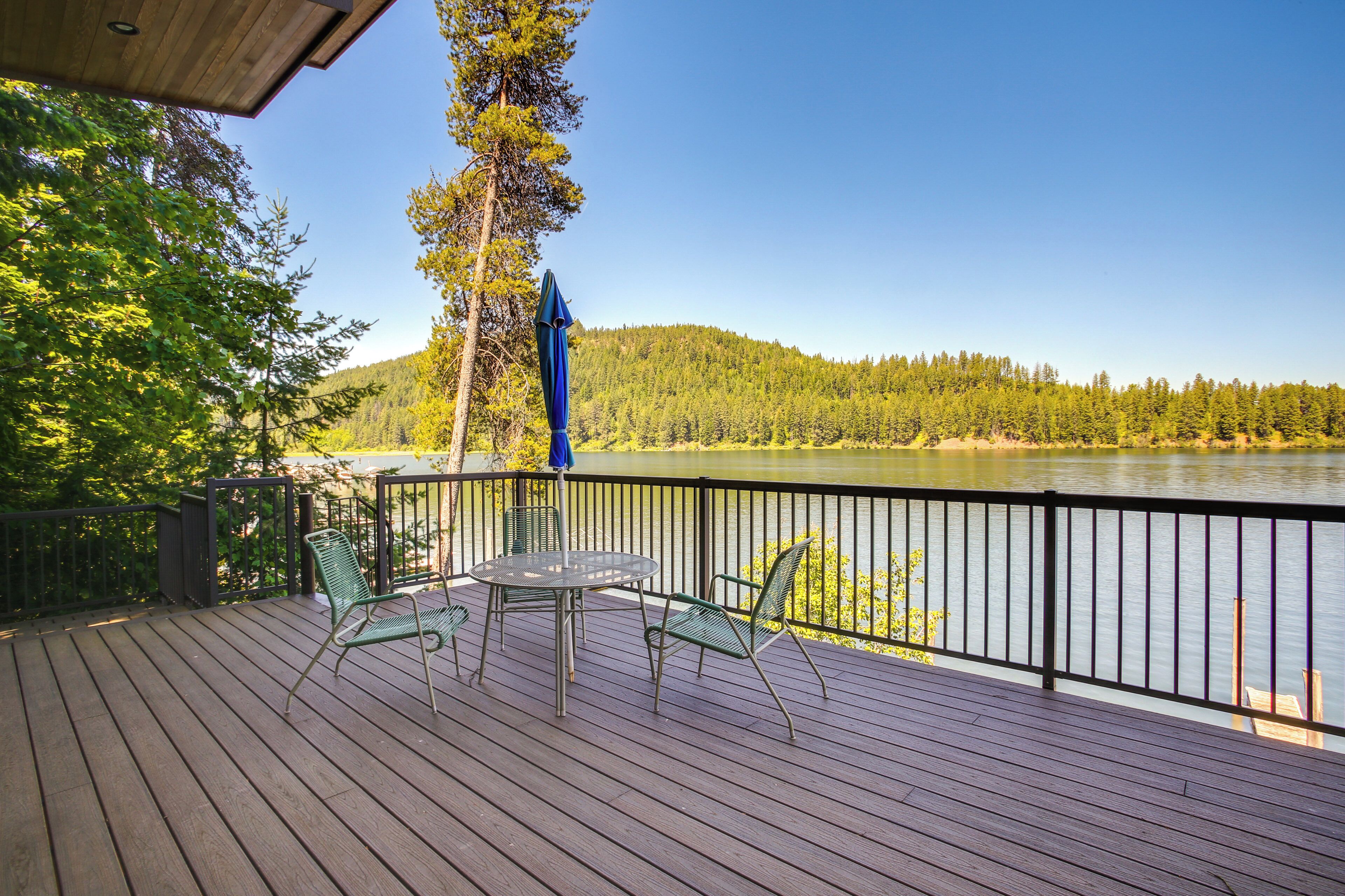 A wooden deck with a table and chairs, overlooking a lake with a forested hillside in the background.