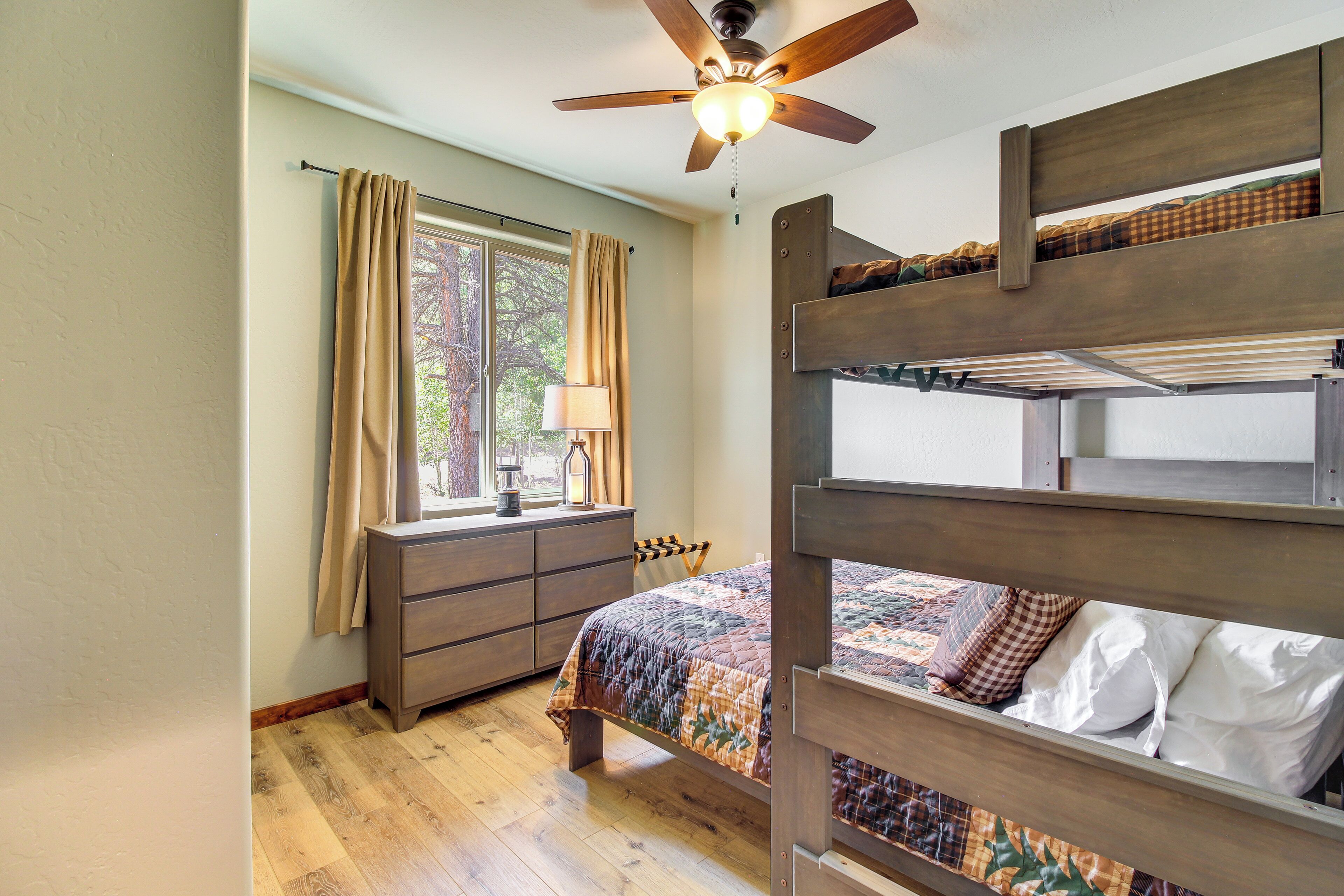 Une chambre avec un lit superposé, un ventilateur de plafond, une fenêtre avec des rideaux, une commode et une couvre-lit matelassé.