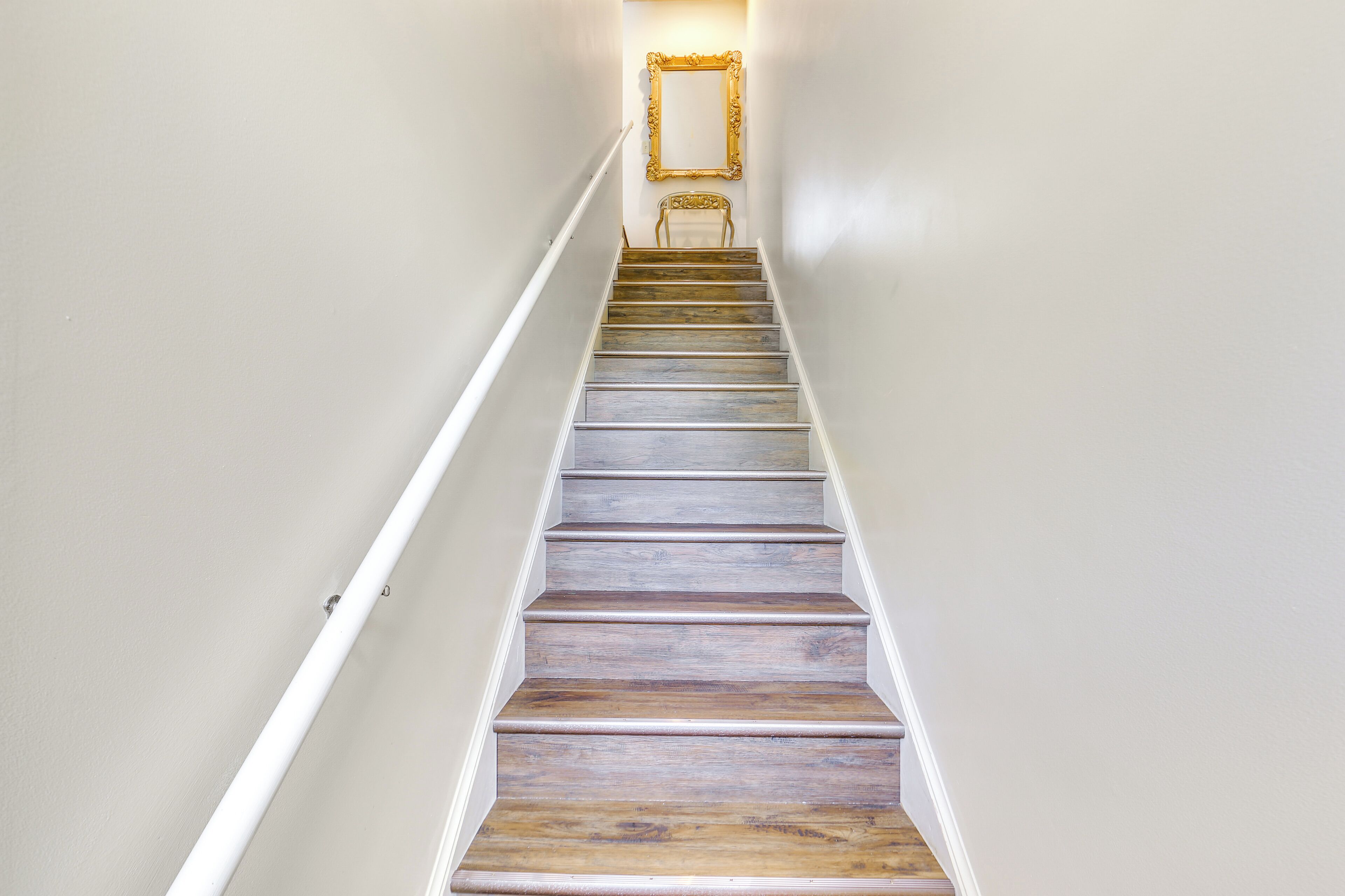 A staircase with wooden steps and a white handrail, leading to a room with a chair and a framed mirror on the wall.