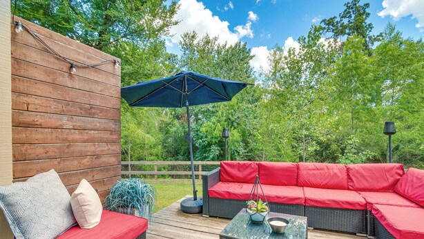 Une terrasse extérieure avec un canapé en osier et des coussins rouges, une table en verre avec un saladier, et un parasol bleu.