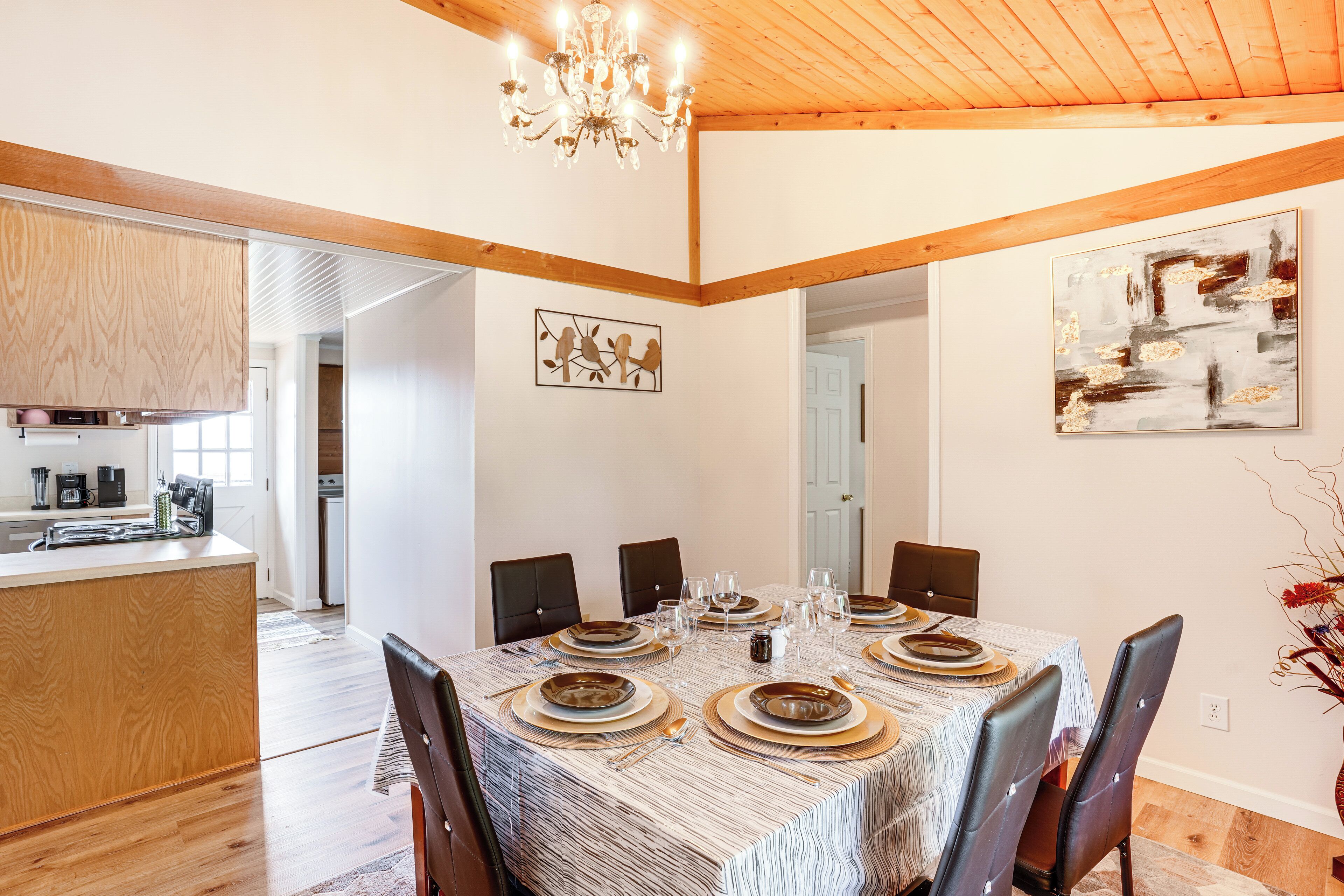 Un coin repas avec une grande table dressée pour six personnes, entourée de chaises, dans une pièce dont le plafond est orné de poutres en bois et qui est illuminée par un lustre.