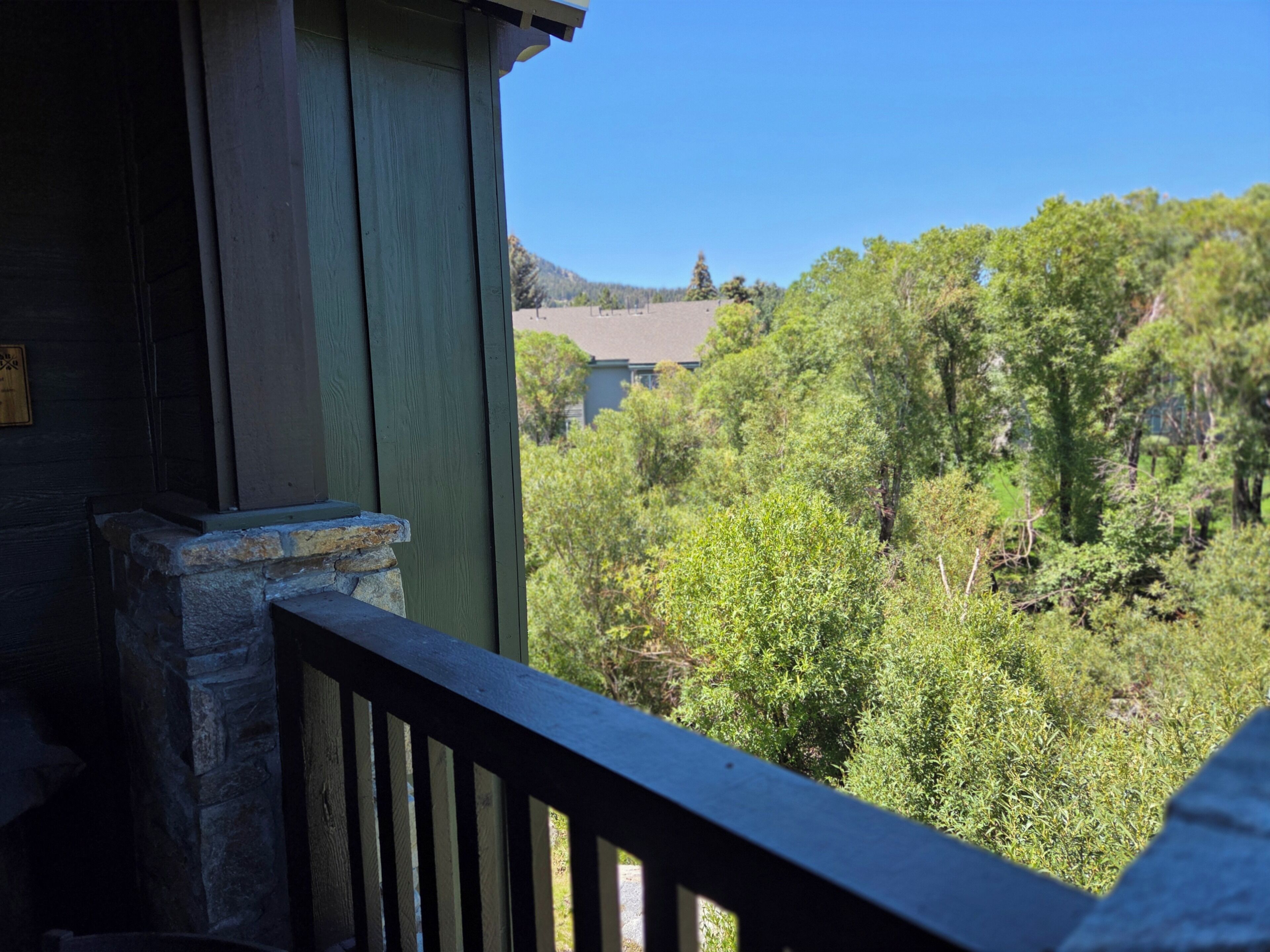 Ein hölzerner Balkon mit Steinunterbau, der einen üppigen, grünen Landschaftsbereich mit Bäumen und einem entfernten Gebäude überblickt.