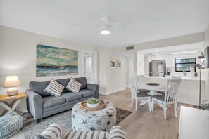 En moderne stue med en grå sofa, en rund skammel med en plante, og et spiseområde med hvide stole og et bord.