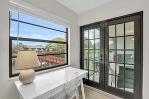 Ein Zimmer mit weißem Schreibtisch, einer Lampe, einem großen Fenster mit Jalousien und einer Schiebeglastür zu einem Außenbereich mit Stühlen und einem Tisch.