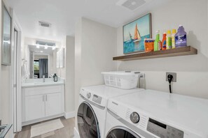 A laundry room with a Samsung washing machine, a dryer, a shelf with cleaning supplies, and a painting of sailboats. - E-234 -Deluxe Updated Condo With Beautiful Views! (Naples)