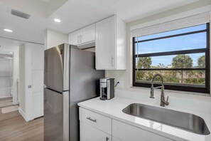 A modern kitchen with white cabinetry, a stainless steel refrigerator, a coffee maker, and a large window with a view of palm trees. - E-234 -Deluxe Updated Condo With Beautiful Views! (Naples)