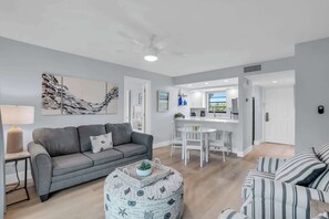 A modern living room with a grey sofa, a striped couch, and a round ottoman with a plant. A white ceiling fan and a kitchen area with a white countertop and chairs are visible. - F-237 - Deluxe Condo - Amazing Views and Sleeps 6 (Naples)
