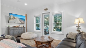 A living room with a large TV, a wooden coffee table, a sofa, and a view of the outdoors through large windows. - Beach Across St | Pool + Hot Tub | Sleeps 12 (Gulf Shores)