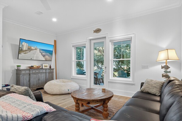 A living room with a large TV, a wooden coffee table, a sofa, and a view of the outdoors through large windows. - Beach Across St | Pool + Hot Tub | Sleeps 12 (Gulf Shores)
