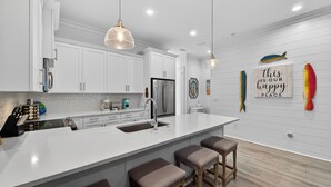 A modern kitchen with white cabinetry, a large island with stools, pendant lighting, and decorative fish wall art. - Beach Across St | Pool + Hot Tub | Sleeps 12 (Gulf Shores)