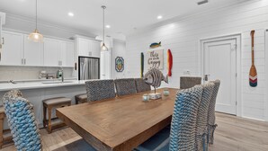 A modern kitchen with white cabinetry, a wooden dining table, and blue patterned chairs. - Beach Across St | Pool + Hot Tub | Sleeps 12 (Gulf Shores)