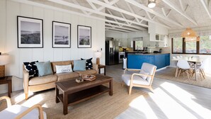 Een moderne woonkamer met een bank, salontafel en stoelen, een keuken op de achtergrond en ingelijste foto's aan de muur.