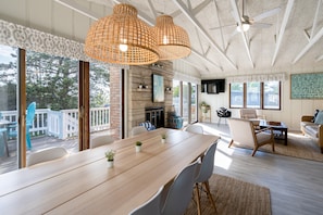 Una habitación amplia con una gran mesa de madera, sillas blancas, chimenea y ventanas grandes que ofrecen vistas al exterior.