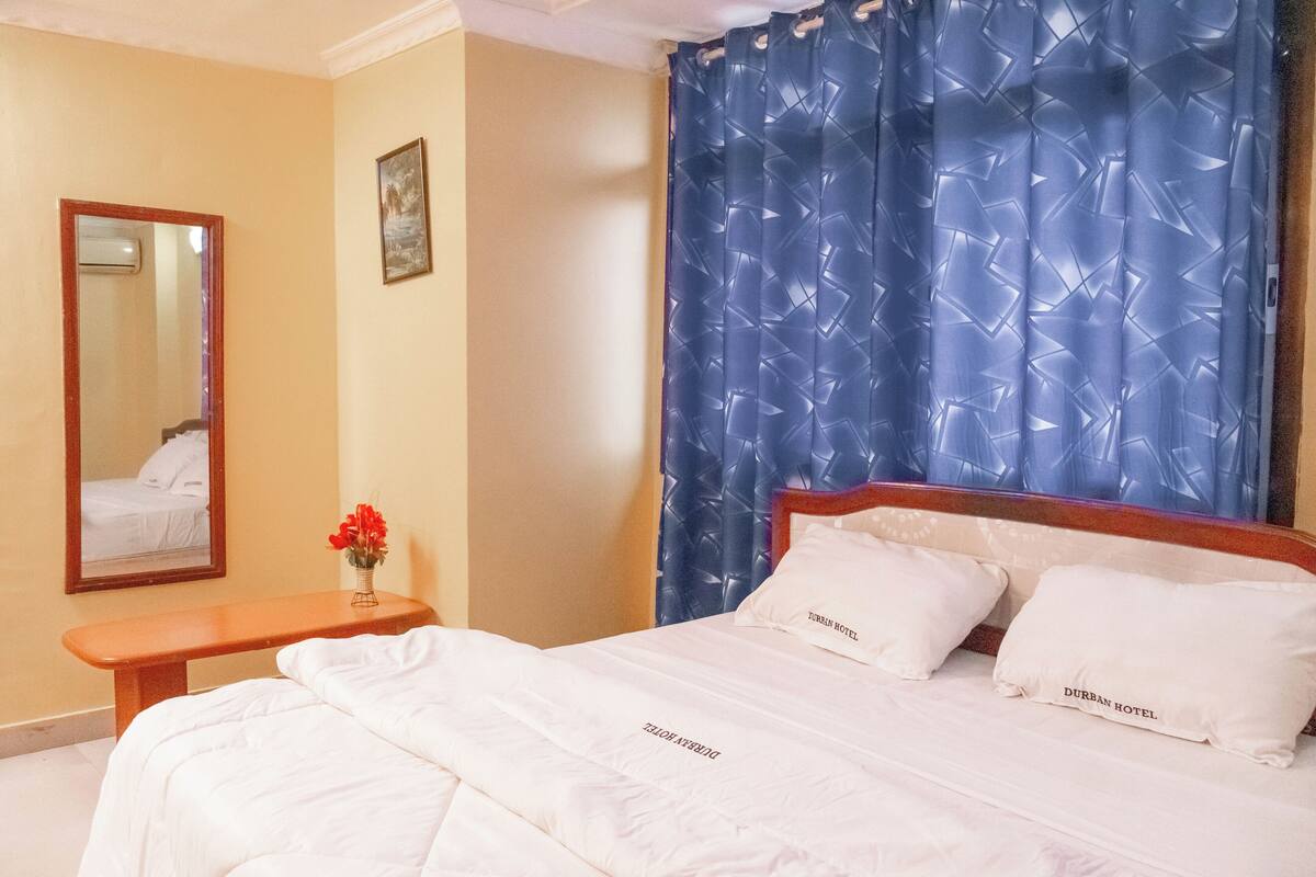 Een hotelkamer met een bed, een nachtkastje met een bloemenvaas, een spiegel en een blauw gordijn.