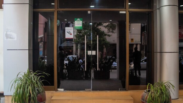 Entrada de un edificio con puertas de vidrio, un letrero verde y dos plantas en macetas.