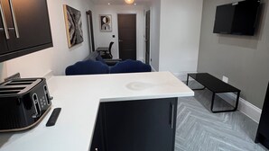 A modern kitchen with a white countertop, black cabinets, and a built-in toaster. There is a bench and a chair in the dining area, and a flat-screen TV mounted on the wall. - Stylish Flat in London, sleeps 4
 (Hornchurch)