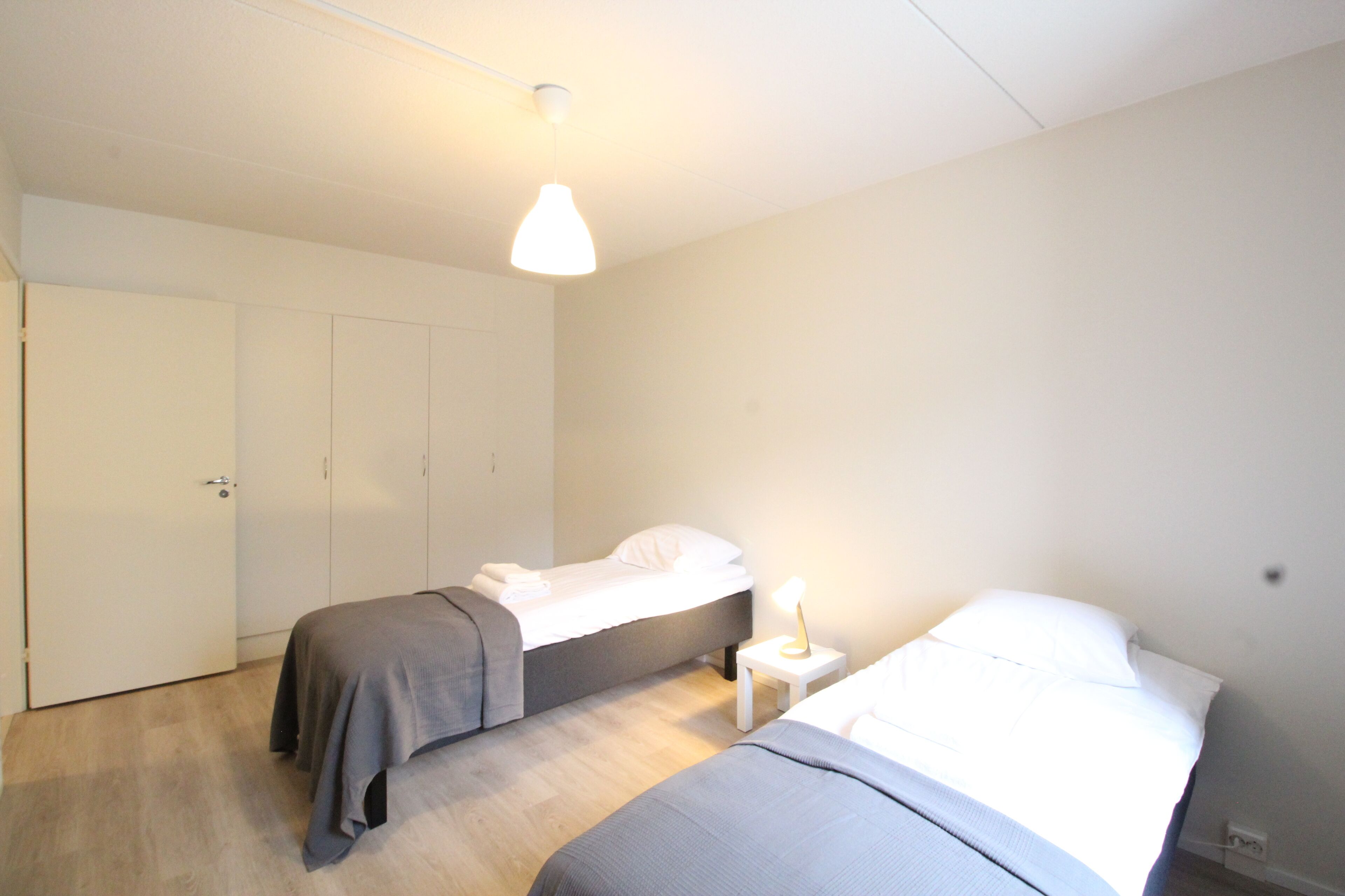 A bedroom with two beds, a white wardrobe, and a bedside table with a lamp.