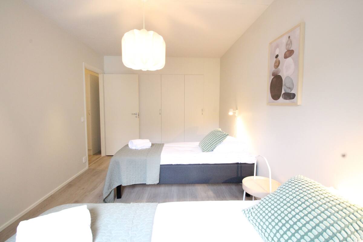 A bedroom with two beds, a bedside table, a chair, and a hanging light fixture.