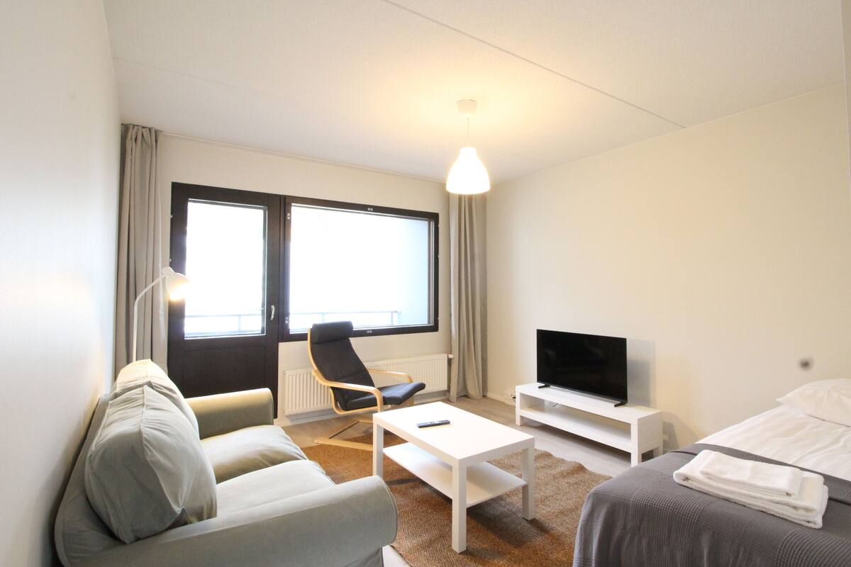 A modern hotel room with a sofa, a TV on a stand, a bed, and a window with curtains.