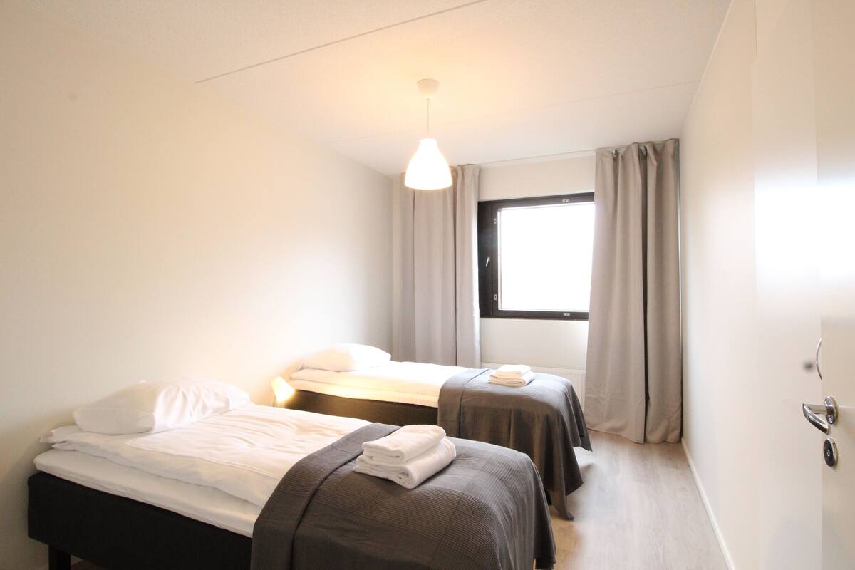 A hotel room with two beds, a window with curtains, and a hanging light fixture.