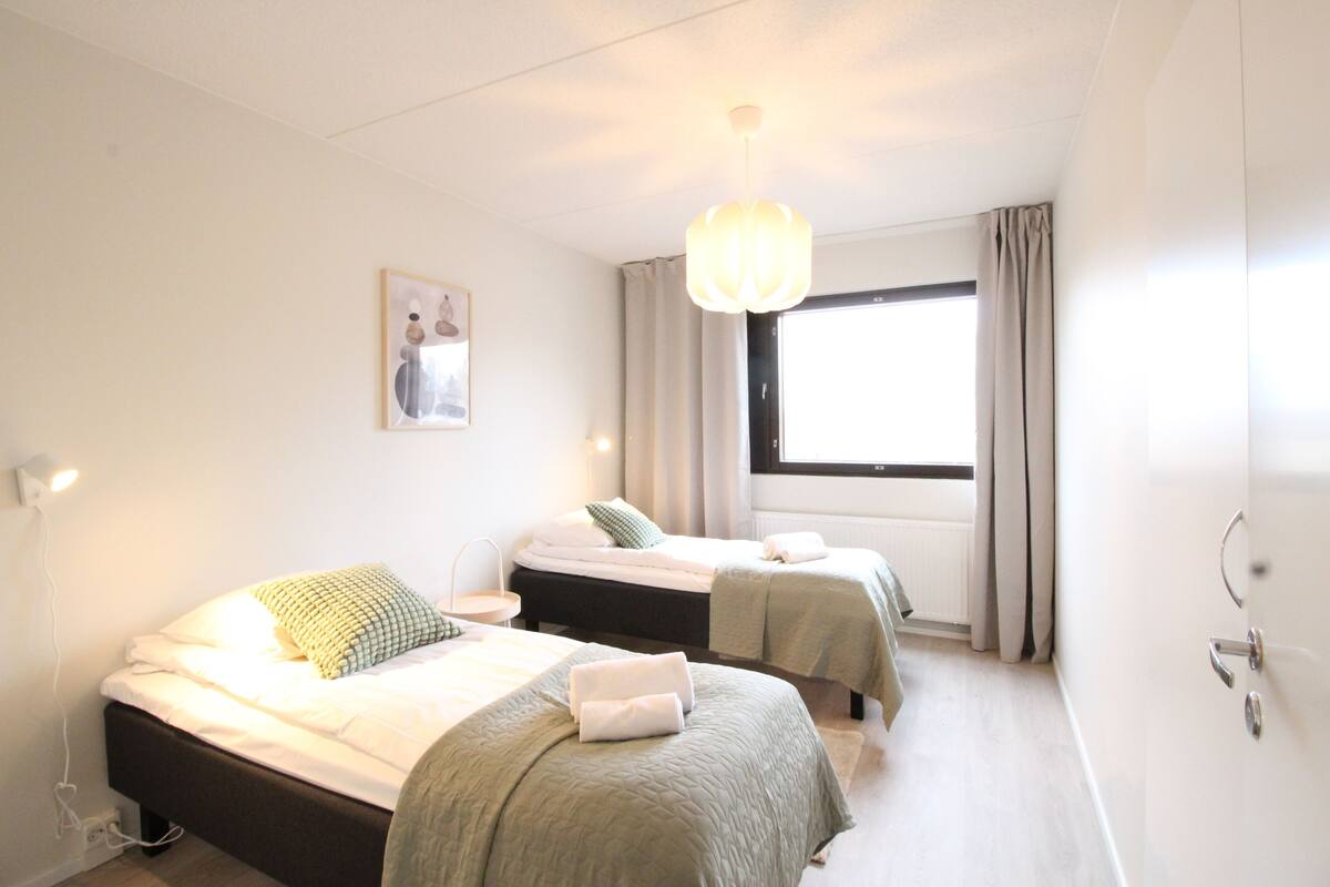 A hotel room with two beds, a large window with curtains, a hanging light fixture, and a wall-mounted picture.