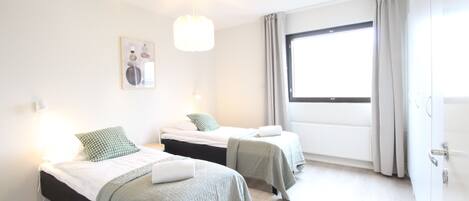 Moderni hotellihuone, jossa on kaksi sänkyä, suuri ikkuna verhoilla, riippuvalaisin ja seinällä oleva taulu.