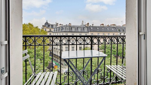 Un balcon orné d’une balustrade en fer forgé, une petite table et une vue sur une rangée d’immeubles à étages multiples, chacun doté de balcons.