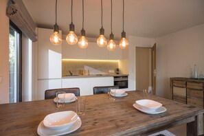 Een moderne eethoek met een houten tafel gedekt voor vier personen, hangende verlichting en een keuken op de achtergrond.