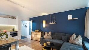 Un salon moderne avec un canapé gris, une petite table ronde et un coin cuisine doté de meubles blancs et d’un plan de travail en bois.