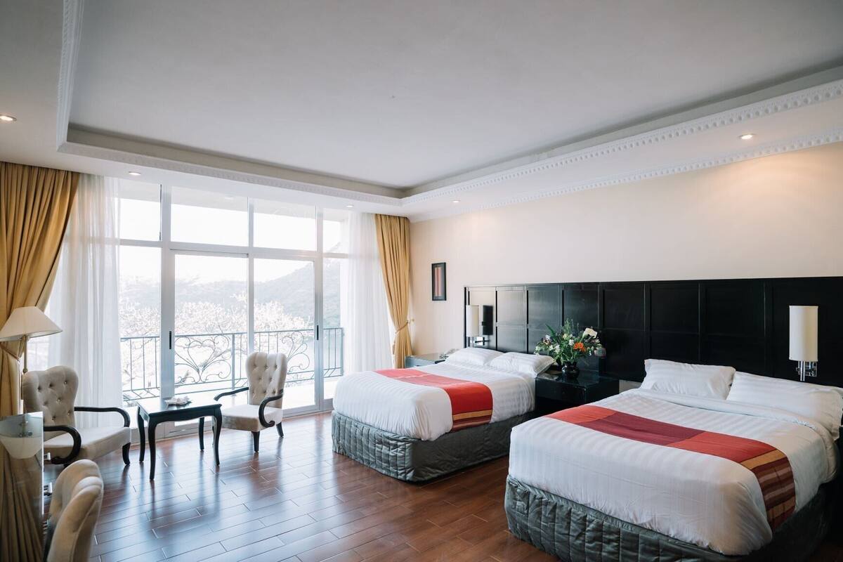 Habitación de hotel con dos camas, un área de estar con una silla, una mesa y vistas al exterior.