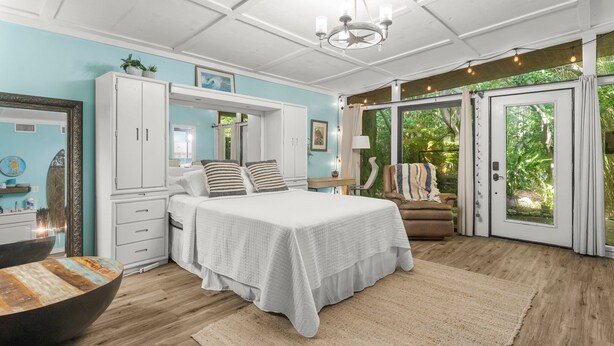A bedroom with a large bed, white wardrobe, and a view of greenery through the glass doors. - Zen Studio at Lotus Garden Cafe (Panama City Beach)