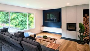 A modern living room with a sectional sofa, a coffee table, a fireplace, and a large window offering a view of greenery. - Creekside Haven Family-Friendly Squamish Home (Squamish)