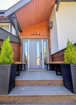 Une entrée moderne, avec une façade en bois, une porte grise et le numéro « 1454 » au-dessus.