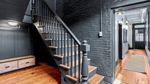 Un escalier avec des marches en bois et des rampes noires, menant à une pièce dont un mur est peint dans une couleur sombre et qui est éclairée par un luminaire suspendu.